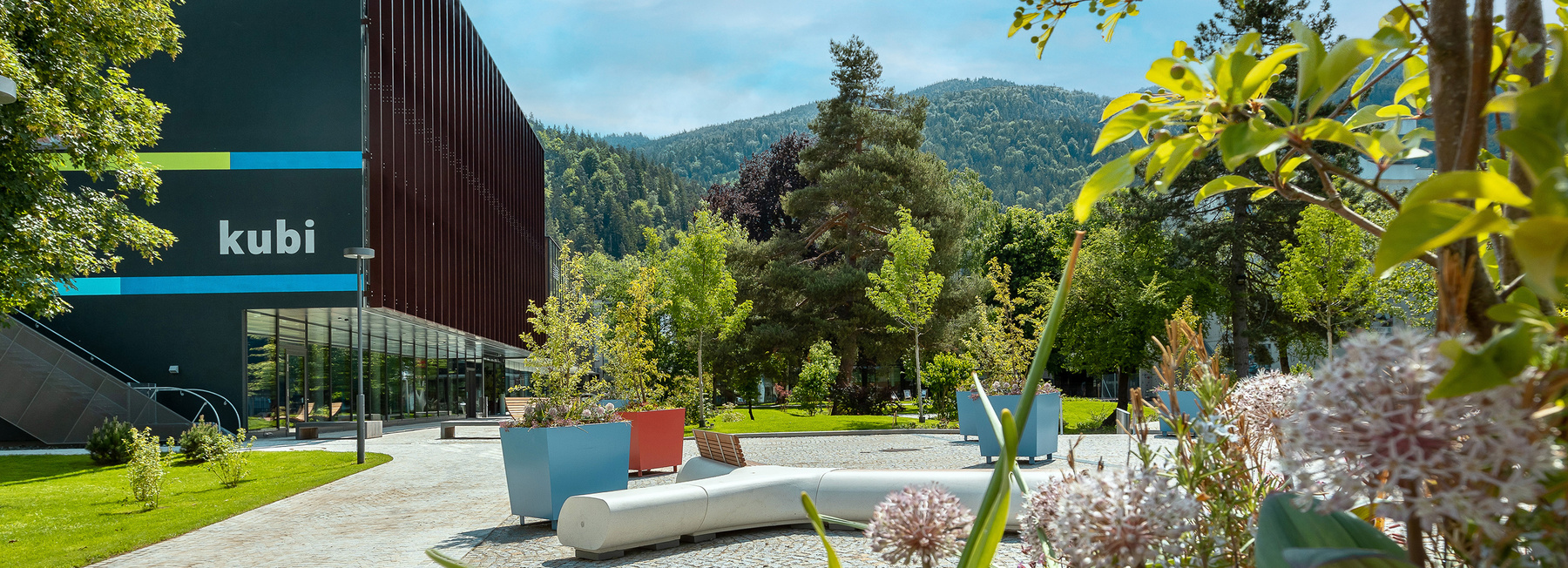 Campus der FH Kufstein Tirol mit kubi-Gebäude, moderner Bau mit Glasfront und dunkler Fassade, davor bunte Sitzmöbel und grüne Bäume im Stadtpark Kufstein, Berge im Hintergrund