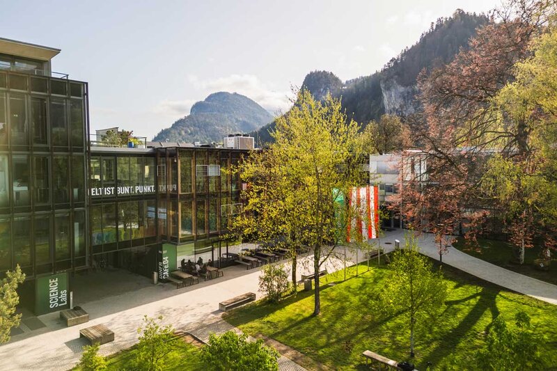 Frühlingshafte Außenaufnahme des Campus der FH Kufstein Tirol mit Glasfassade, Fahnen und Bergkulisse im Hintergrund